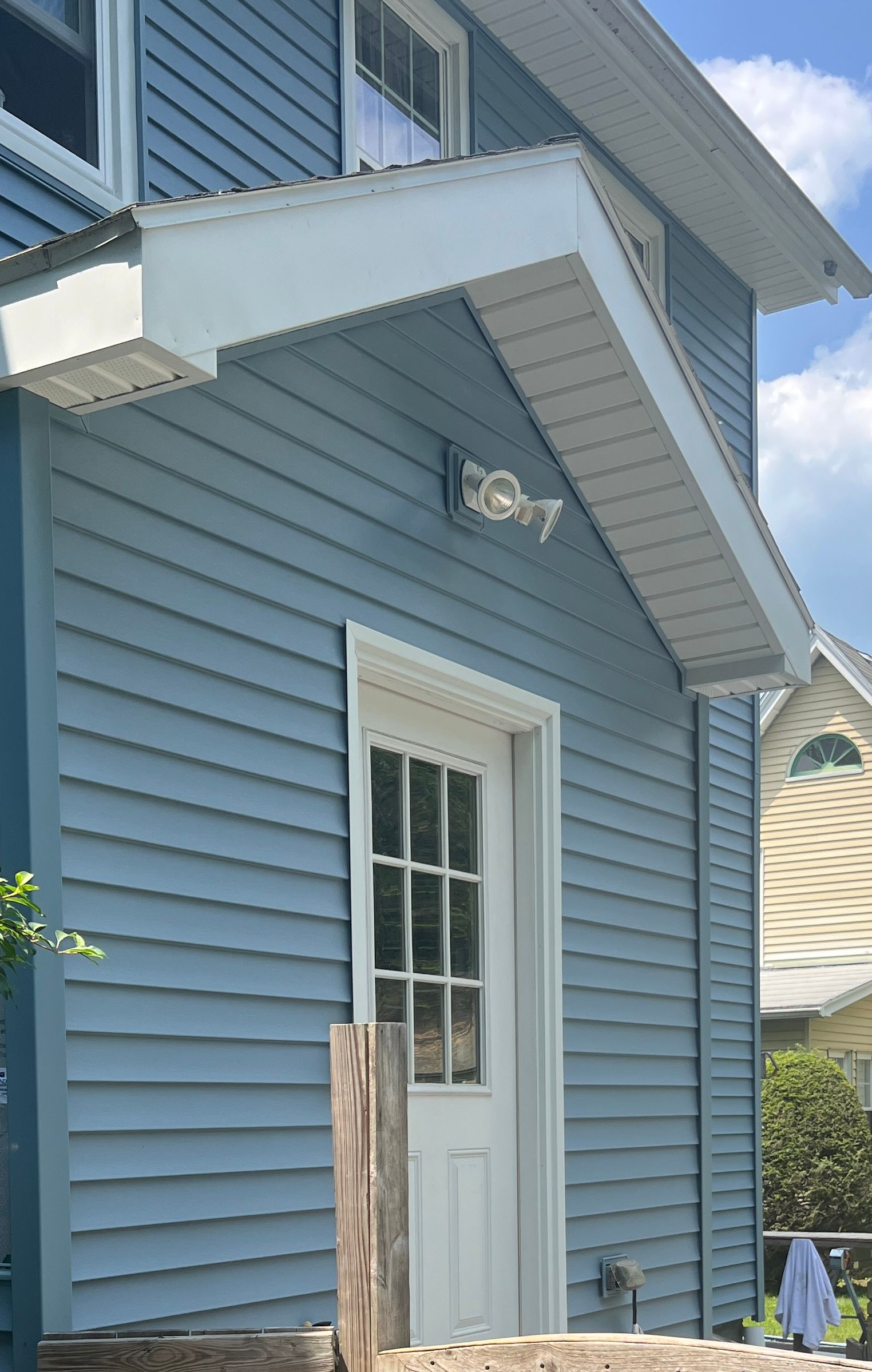 Blue-sided house with white door and awning; bright sunlight.