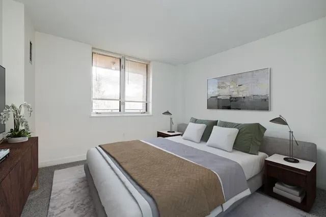 A modern bedroom with a bed featuring a tan and gray spread, bedside tables with lamps, and a landscape wall art piece.