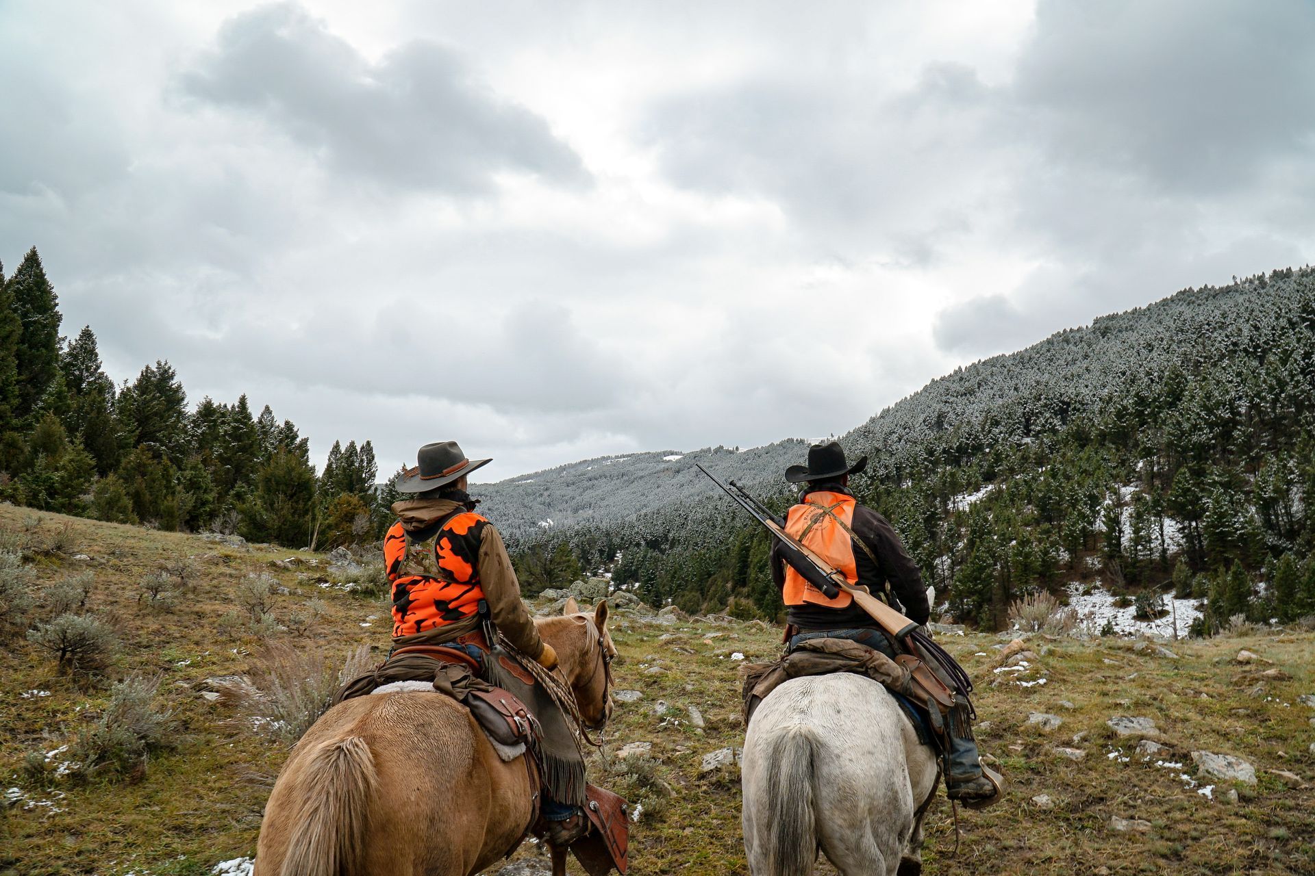 Two men are riding horses in a field with mountains in the background.