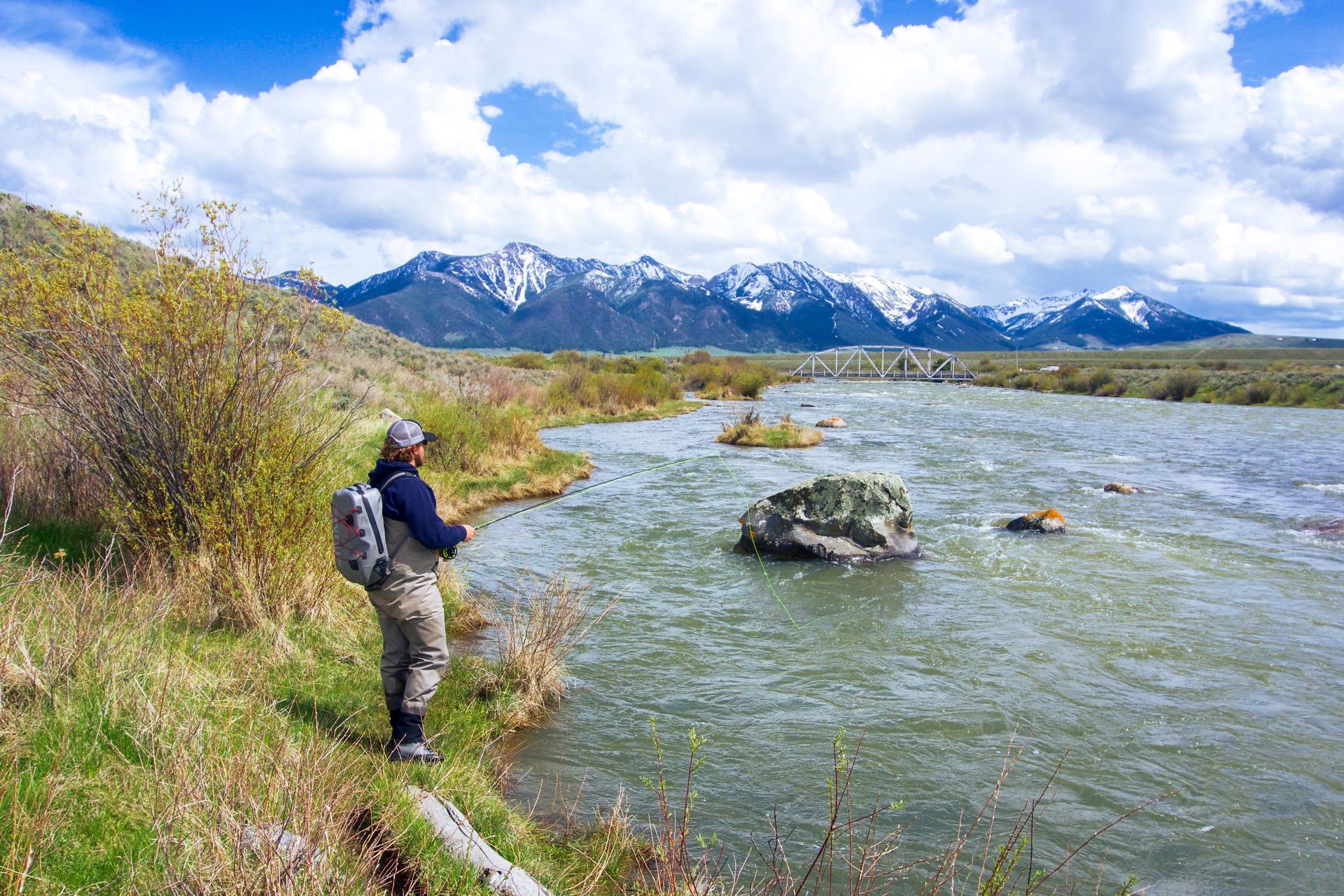 A man is fishing in a river with mountains in the background.