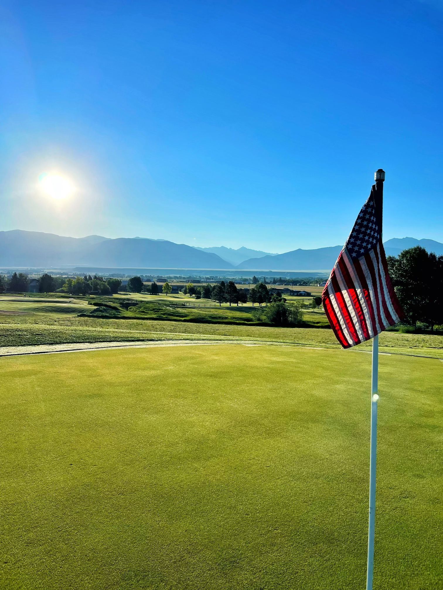 An american flag is flying on a golf course with mountains in the background.