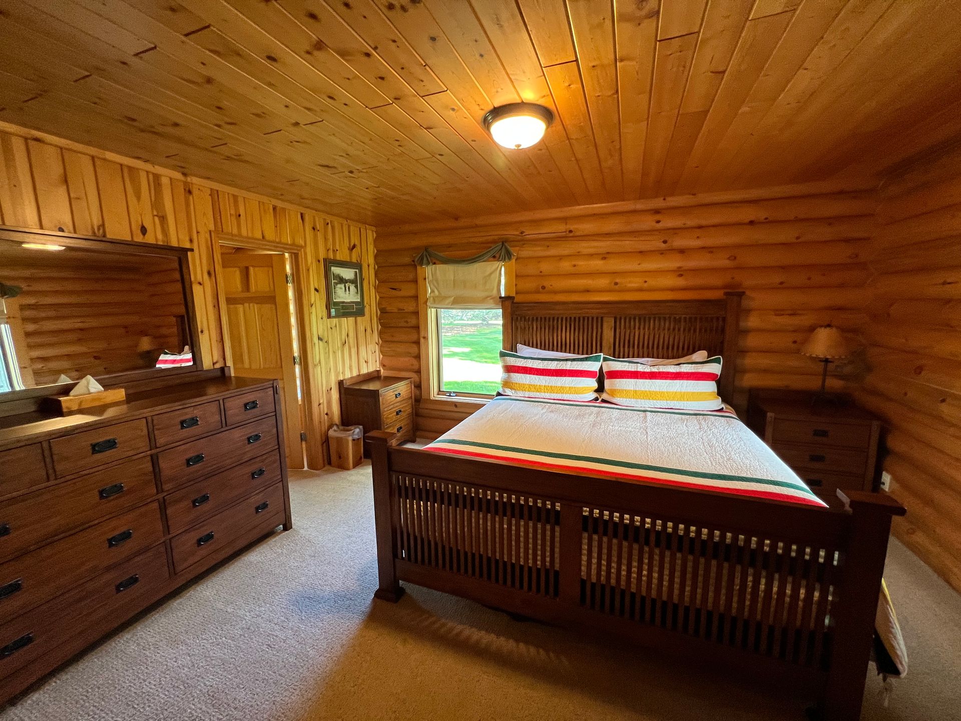 A bedroom in a log cabin with a bed , dresser , mirror and window.