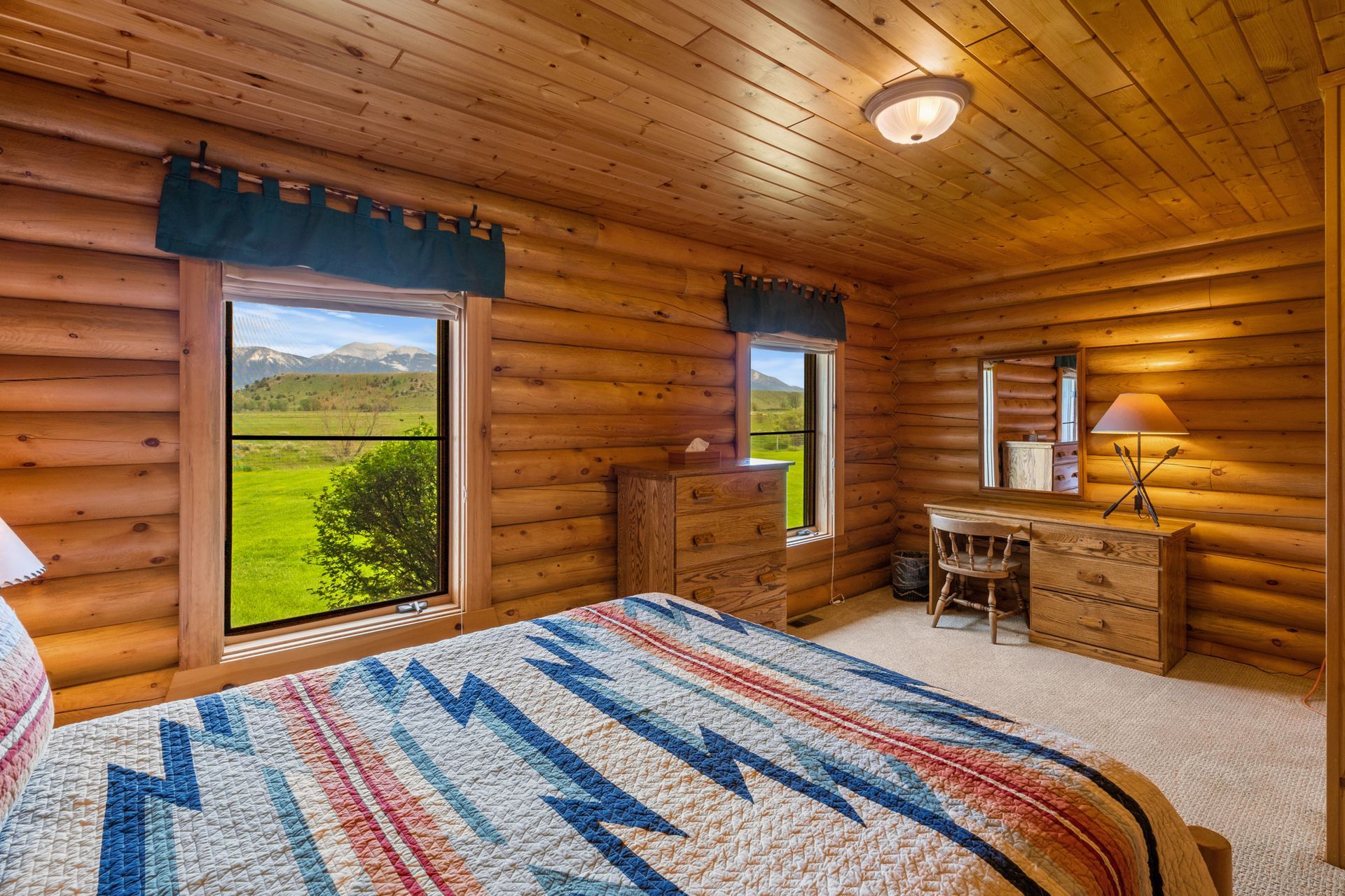 A bedroom in a log cabin with a bed , desk , mirror and window.