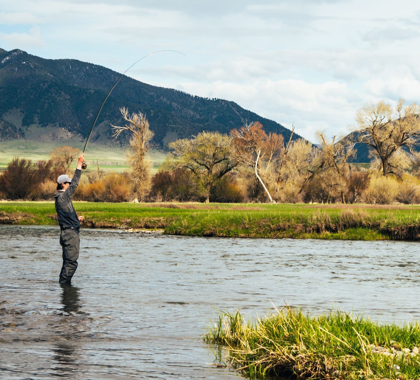 A man is fishing in a river with mountains in the background