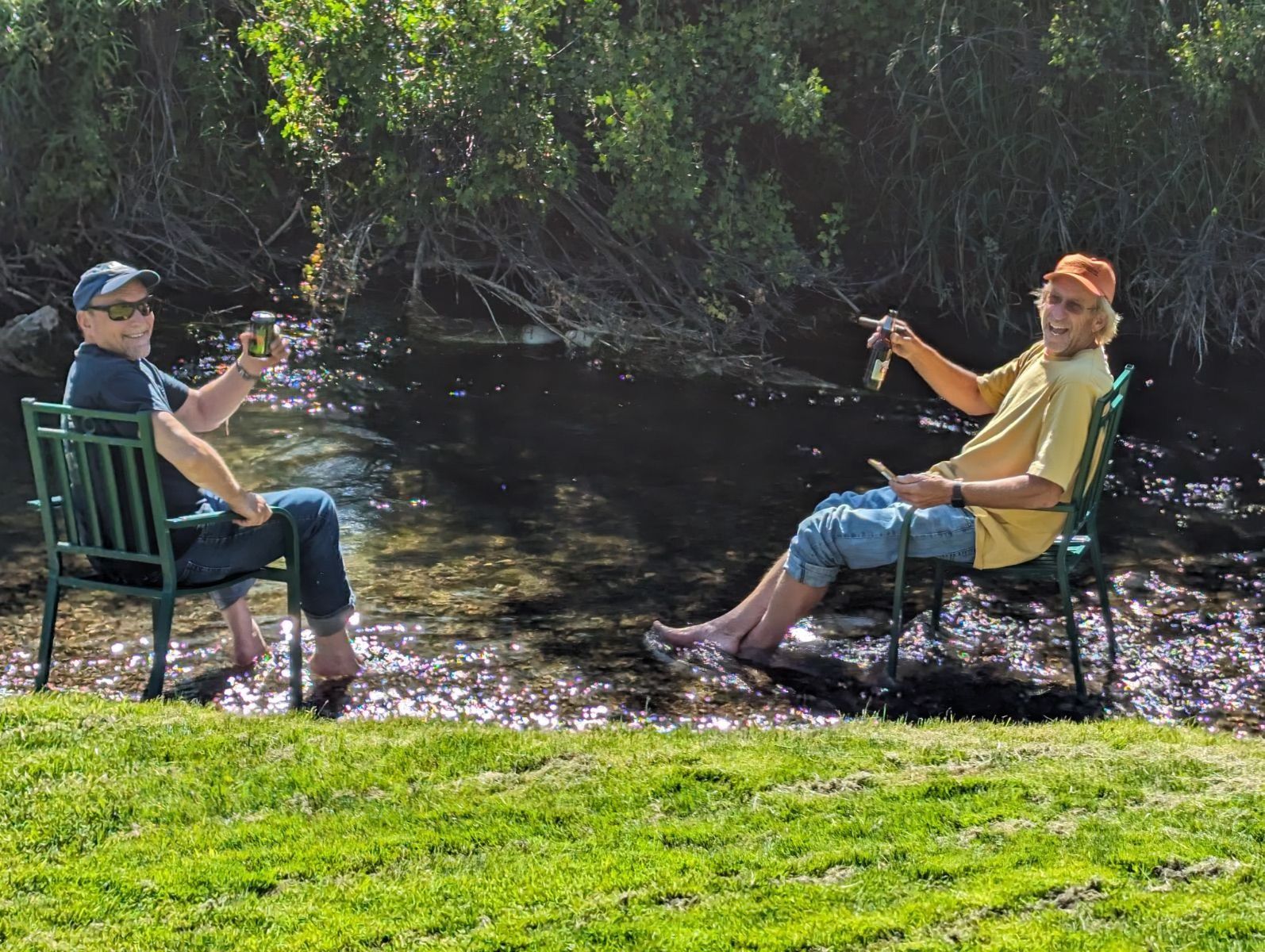 Two men are sitting in chairs near a river drinking beer.