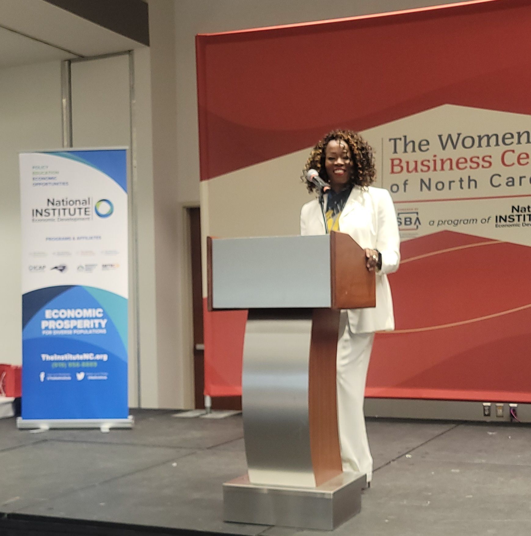 MiMi K stands at a podium in front of a sign that says the women business center of north carolina