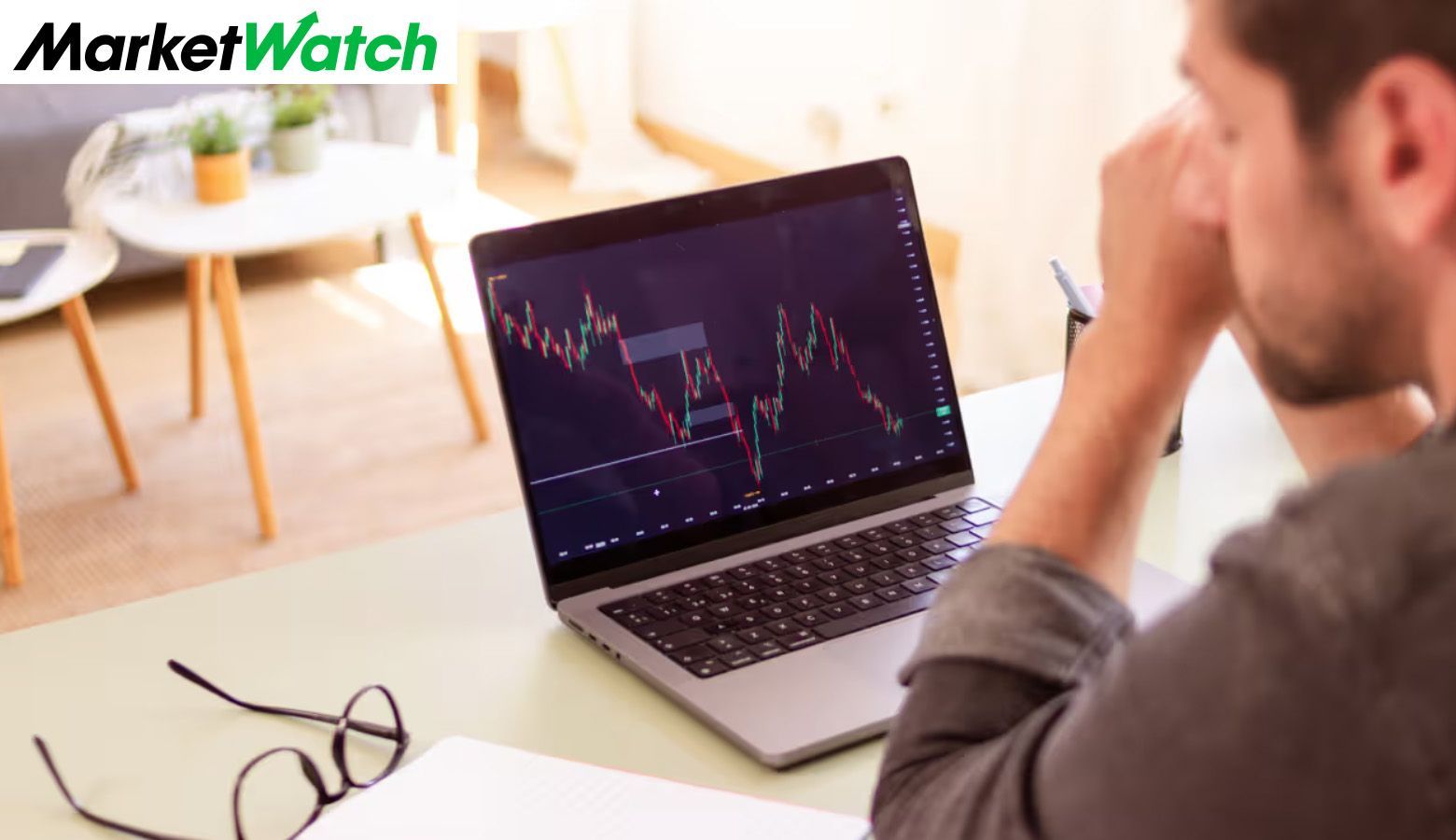 A man is sitting at a desk, looking at stock market graphs on a laptop computer.