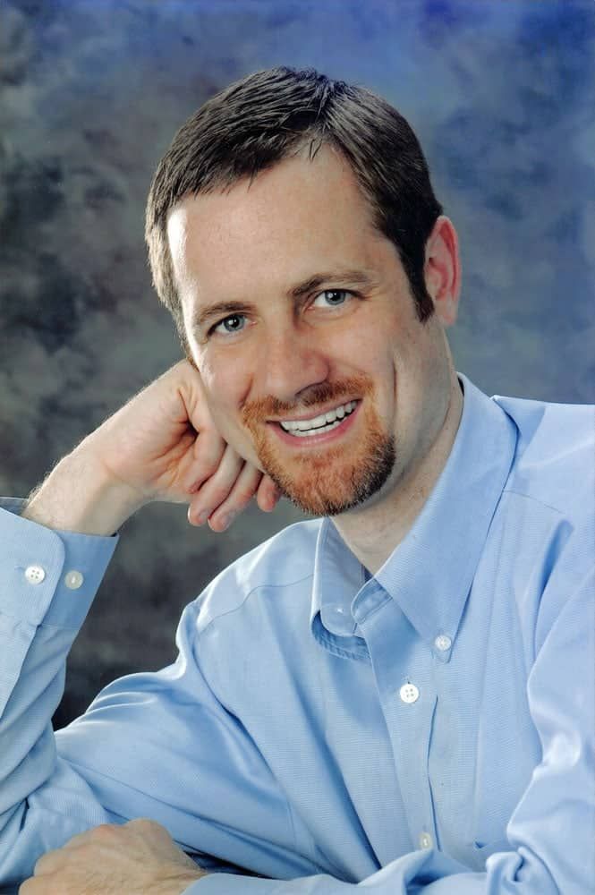 A photo of a man in a blue shirt is smiling with his hand on his chin.