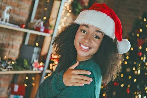 A woman wearing a santa hat and a green sweater is standing in front of a Christmas tree.