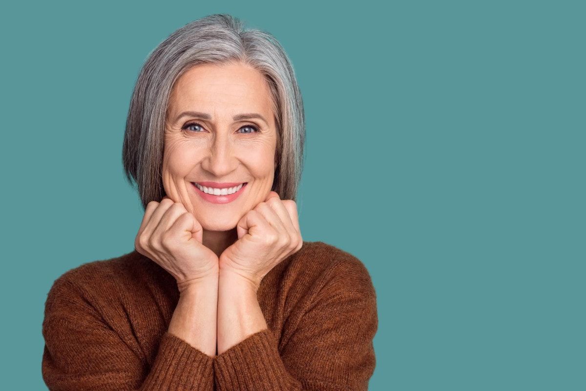 A woman with gray hair is smiling with her hands on her face.