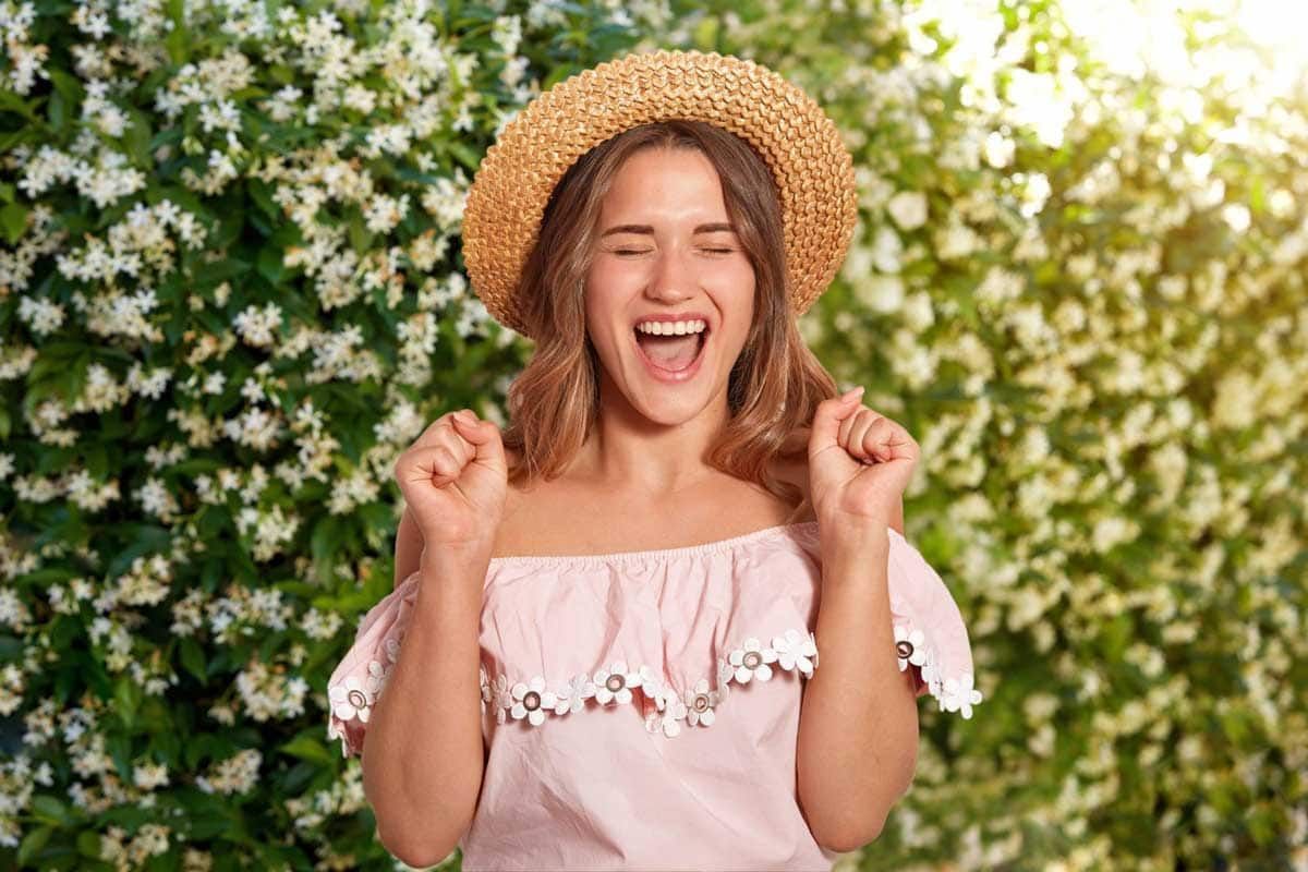 A woman wearing a straw hat and a pink shirt is standing in front of a bush of jasmine flowers.