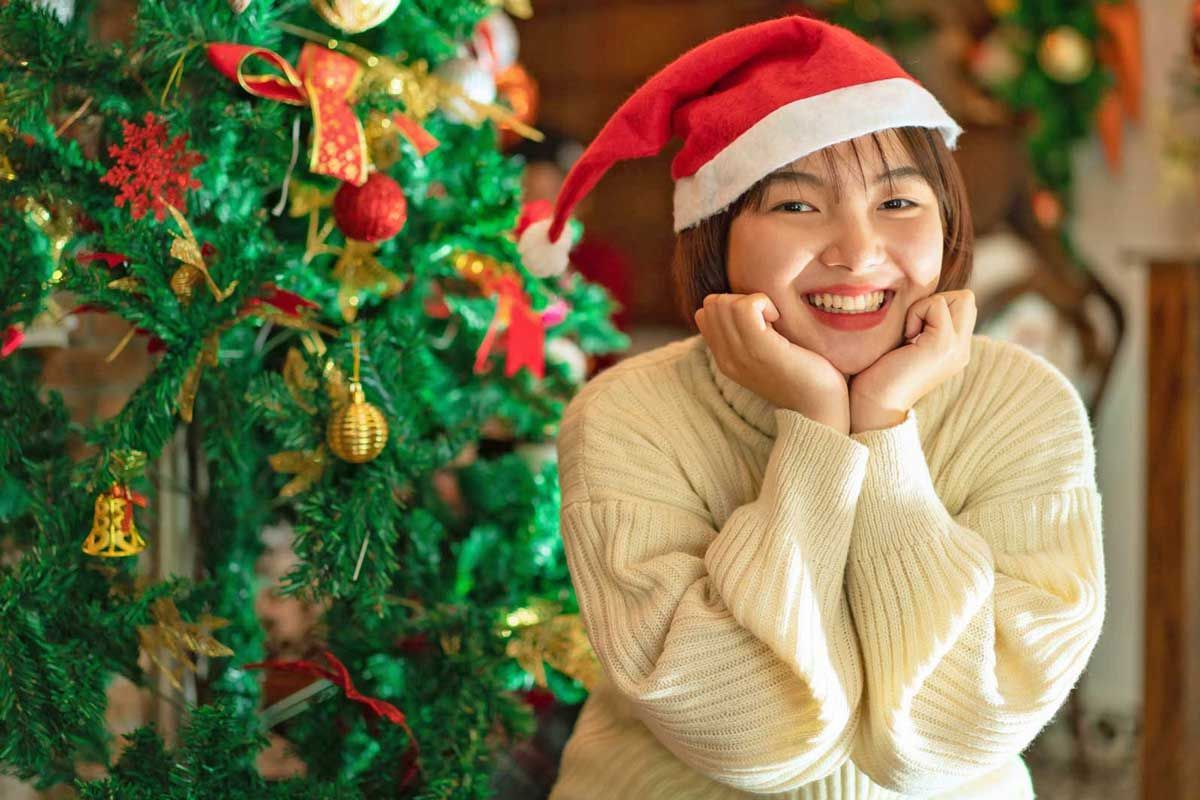 A woman wearing a santa hat is smiling in front of a christmas tree.