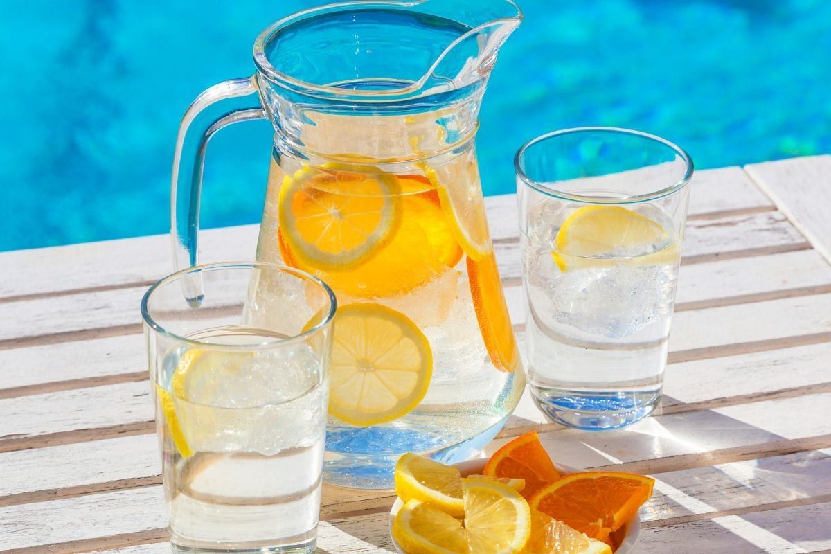 A pitcher of lemonade and two glasses of water on a table next to a pool.