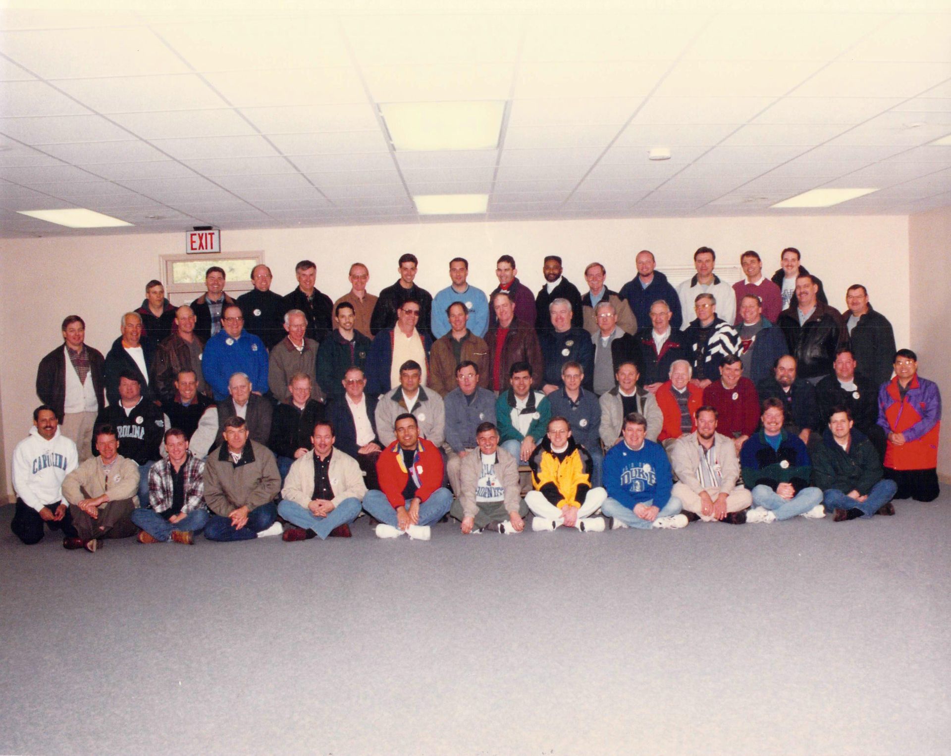 A large group of men pose for a photo in a room with an exit sign on the wall