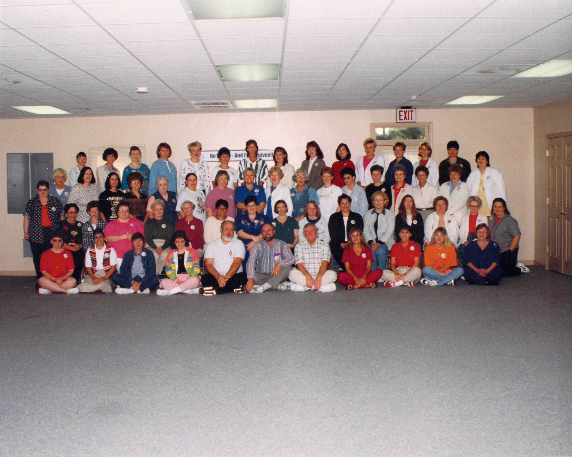 A large group of people are posing for a picture in a room with an exit sign on the wall