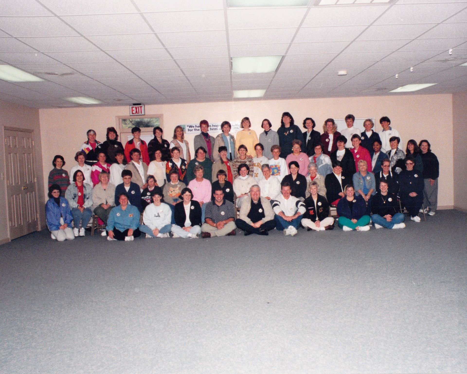 A large group of people are posing for a picture in a room with an exit sign on the wall