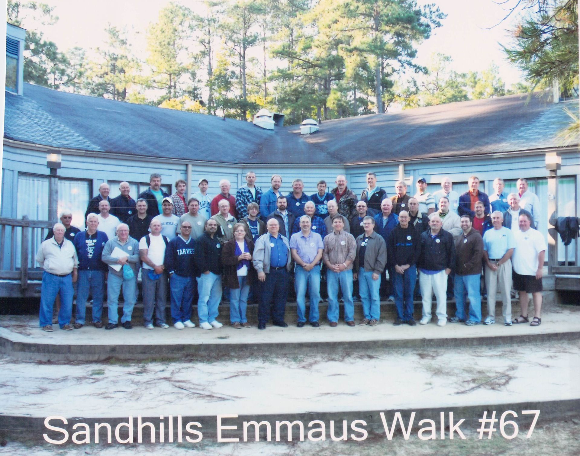 A group of people standing in front of a building that says sandhills emmaus walk # 67
