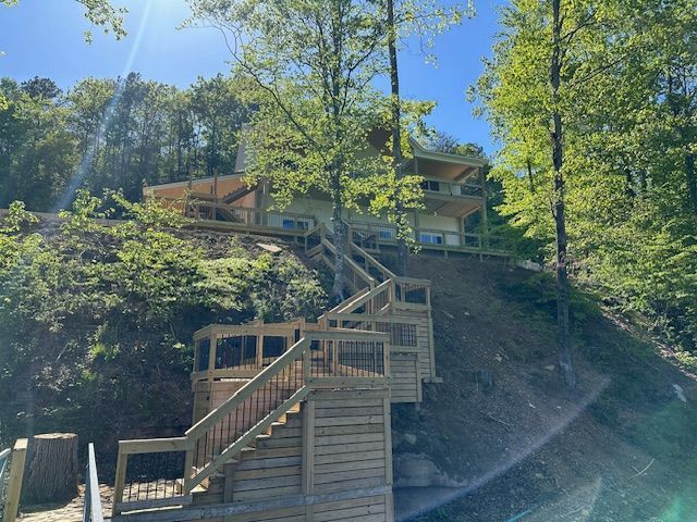 Wooden stairs ascend a hillside to a light-colored house nestled among trees under a blue sky.