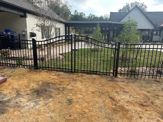 Black metal fence with arched gate in front of a house, set on grass and dirt.