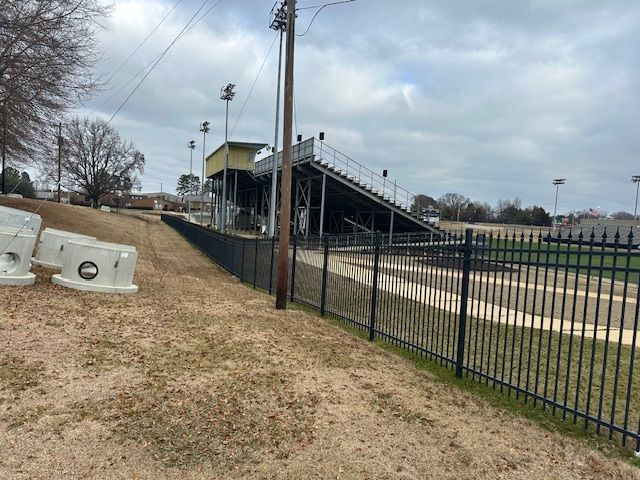 Black stadium with bleachers, lights, and a fence next to a field on a cloudy day.