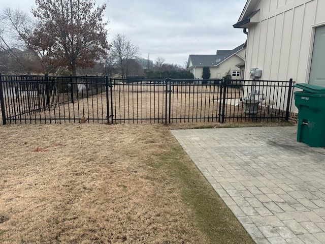 Black metal fence encloses a backyard with brown grass, brick patio, and a house.