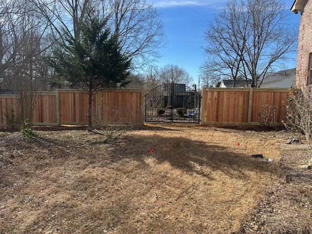 Wooden fence with a gate, in a backyard. Brown grass, bare trees, and a clear, blue sky.
