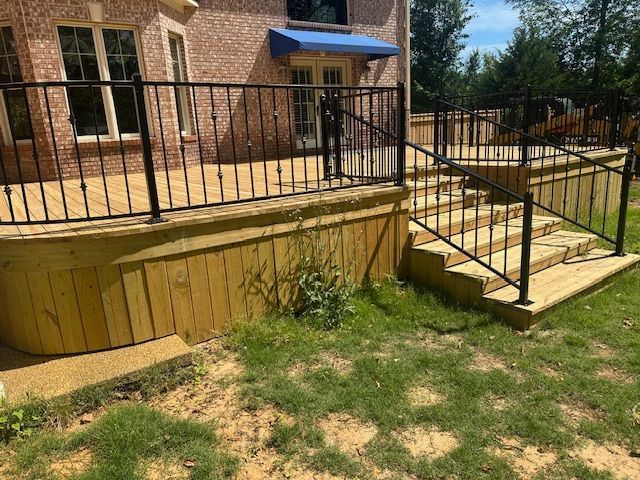 Wooden deck with black railings and steps leading to a house with a brick facade and blue awning.