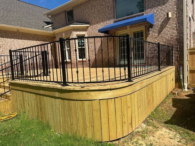A backyard deck with black railing and wooden skirt. Curved edge, green grass, and brick house visible.