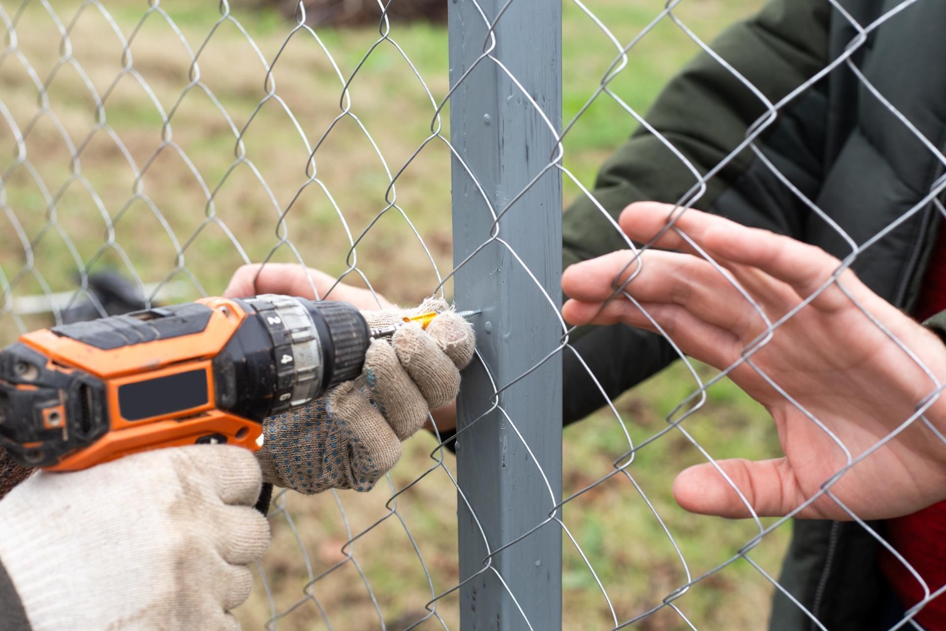 Person using a drill to attach wire mesh fencing to a gray fence post outdoors.
