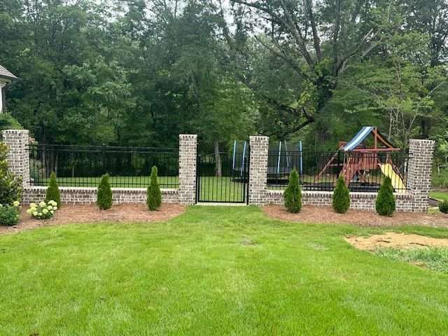 Brick and black metal fence enclosing a grassy yard, gate in the center, playground visible in the background.