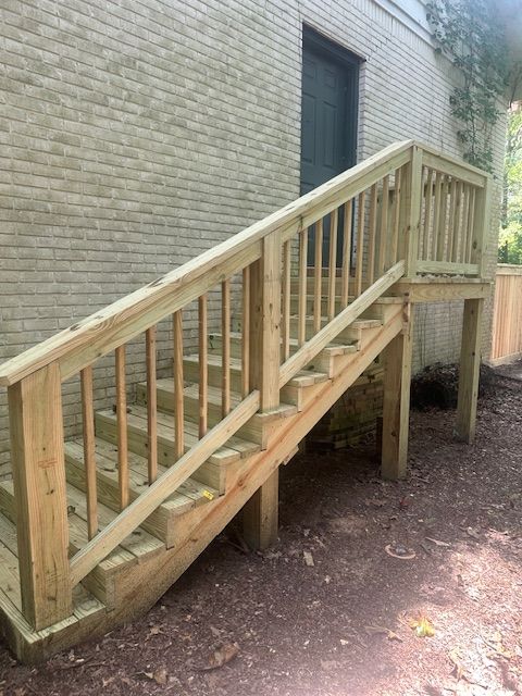 Wooden outdoor staircase with railings, built against a light brick wall, leading up to a small landing.