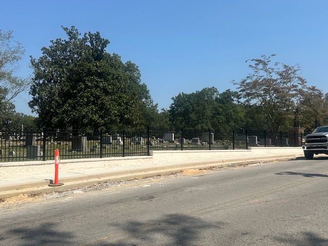 Cemetery behind black fence along a road, trees and sky visible. A car is on the right side.