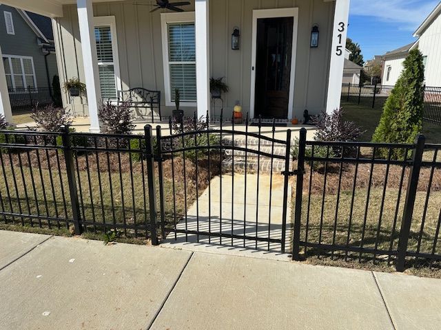 Black metal fence with gate in front of a light-colored house with a sidewalk.