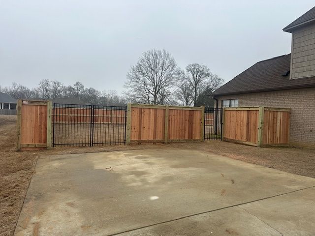 A backyard concrete patio with a wooden fence and metal gate; a brick house is visible.