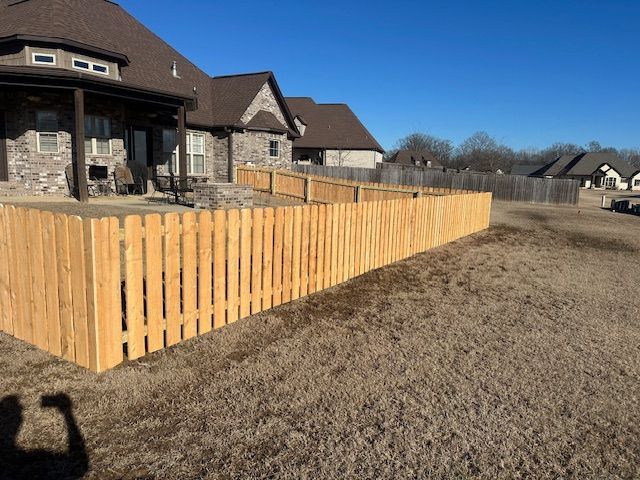 A wooden picket fence surrounds a grassy yard, partially encircling a large house with stone and brick exterior.