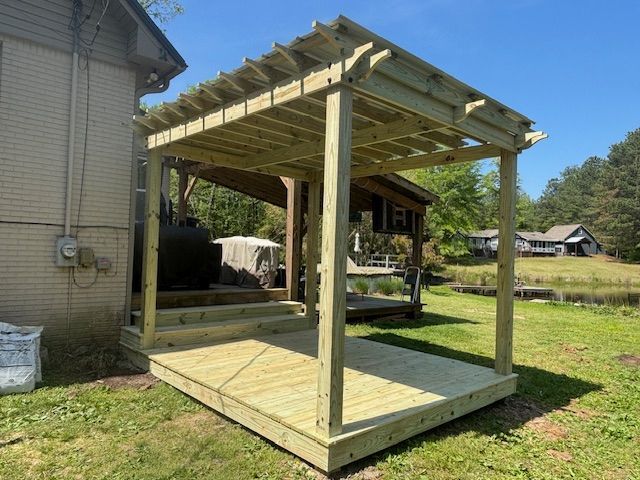 Wooden pergola attached to a brick building. The structure sits on a deck with steps, overlooking a grassy yard.