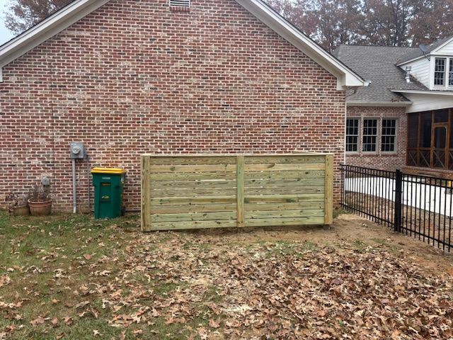 Brick house exterior with a wooden fence concealing trash cans; fallen leaves on the ground.