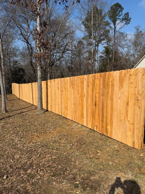 Long, wooden privacy fence in a grassy yard, surrounded by trees under a blue sky.