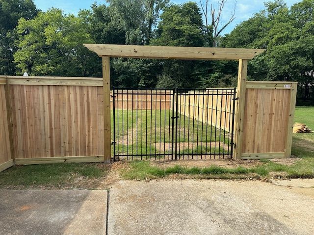Wooden fence with black wrought-iron gate and pergola-style top, set in a grassy yard.