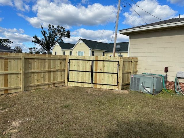 Wooden fence with black metal gate in grassy yard. A/C unit and tan building. Blue sky with clouds.