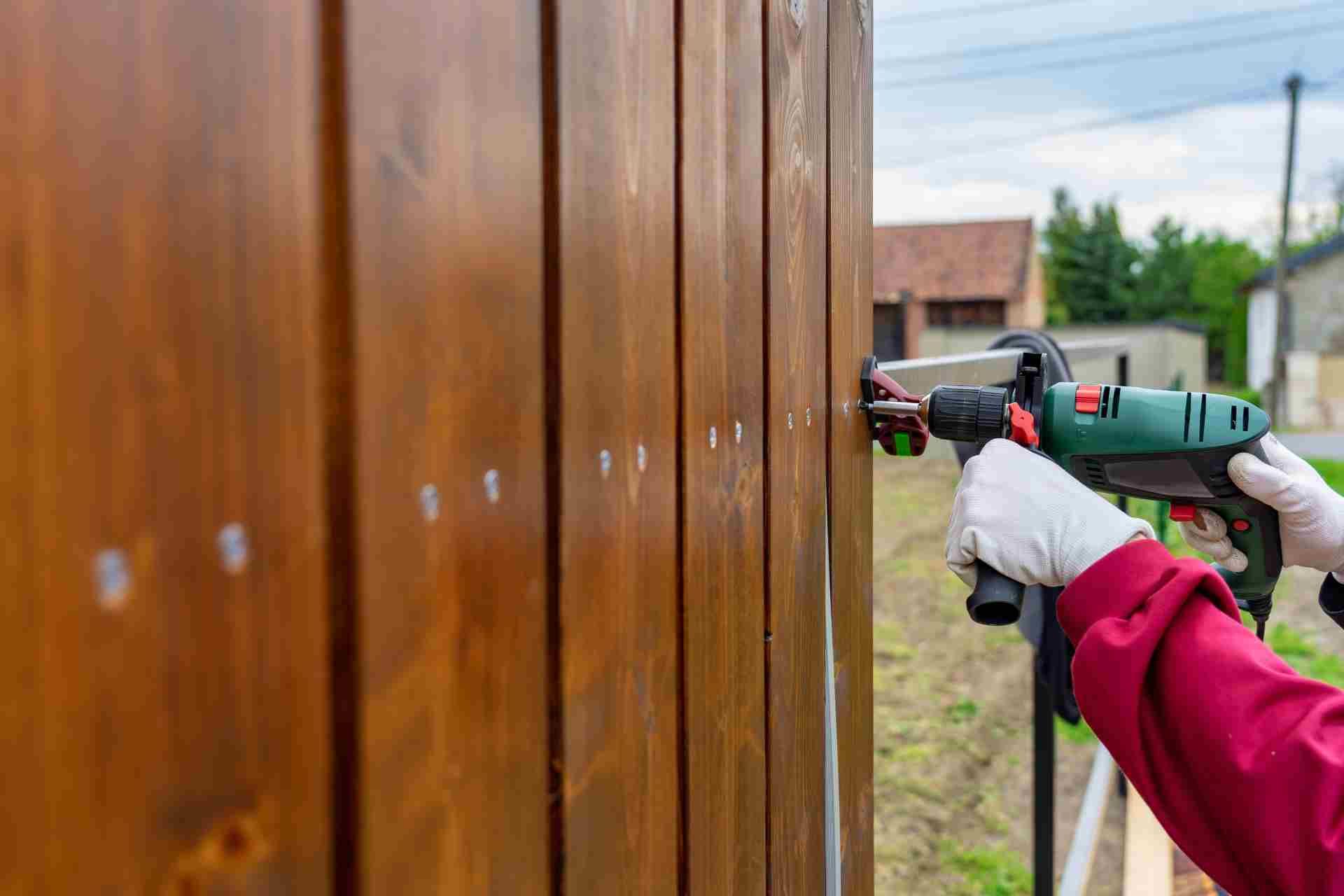 Person in red shirt and gloves uses a drill on a wooden fence outdoors.