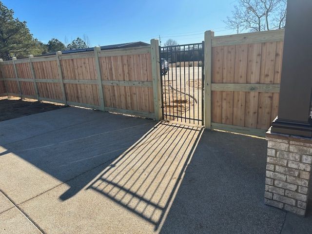 Wooden fence with a black metal gate on a concrete patio; sunny outdoor setting.