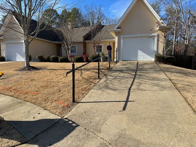 A house with a driveway and a black fence. A person stands near the house. Dry, brown grass.