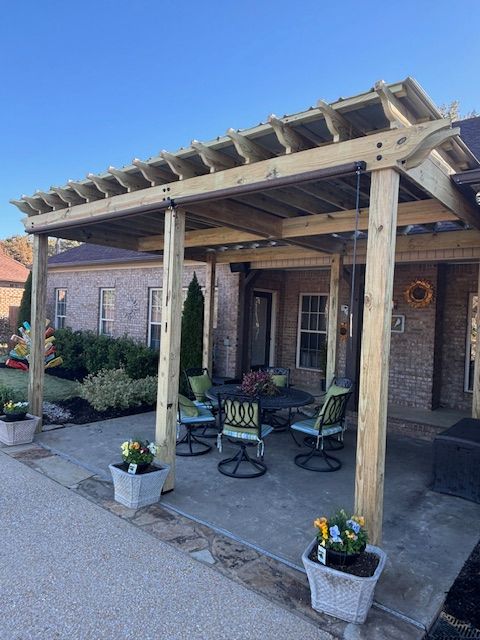 Wooden pergola over a patio with chairs and potted flowers; house in background.