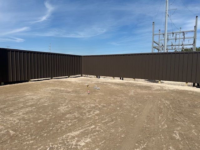 Brown metal fence surrounds a sandy area under a blue sky, with utility structures visible in the distance.
