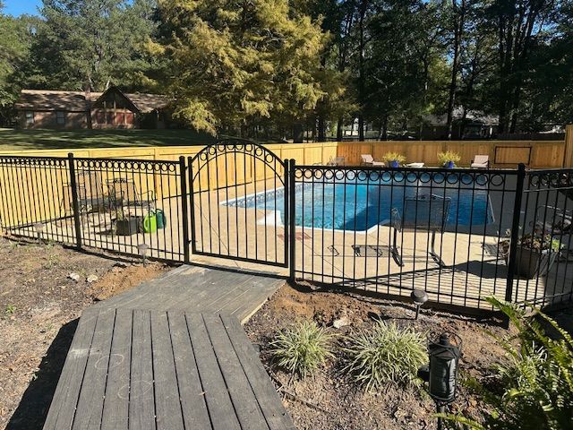 Black iron fence with gate surrounds a swimming pool. Wooden walkway leads to the gate.