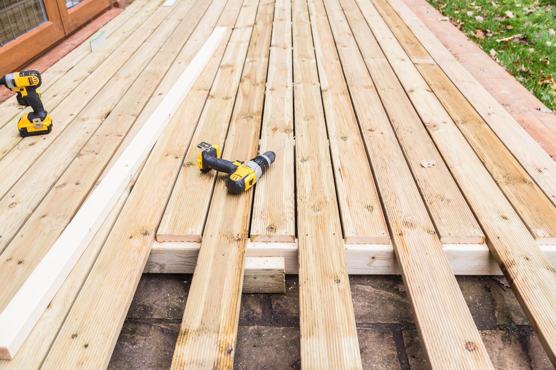 Wooden deck construction: light-colored planks, power drills, and wood supports in an outdoor setting.
