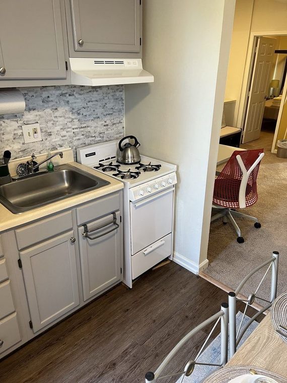 Small kitchen with gray cabinets, white stove and range hood. A glimpse into an adjacent room shows a desk and red chair.