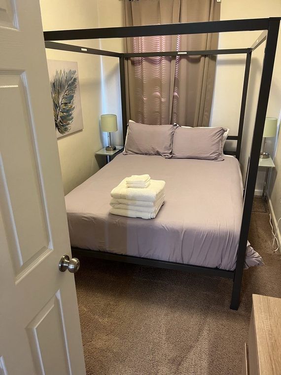 Bedroom with a gray metal canopy bed, lavender bedding, and white towels stacked on top. Two gray nightstands flank the bed.