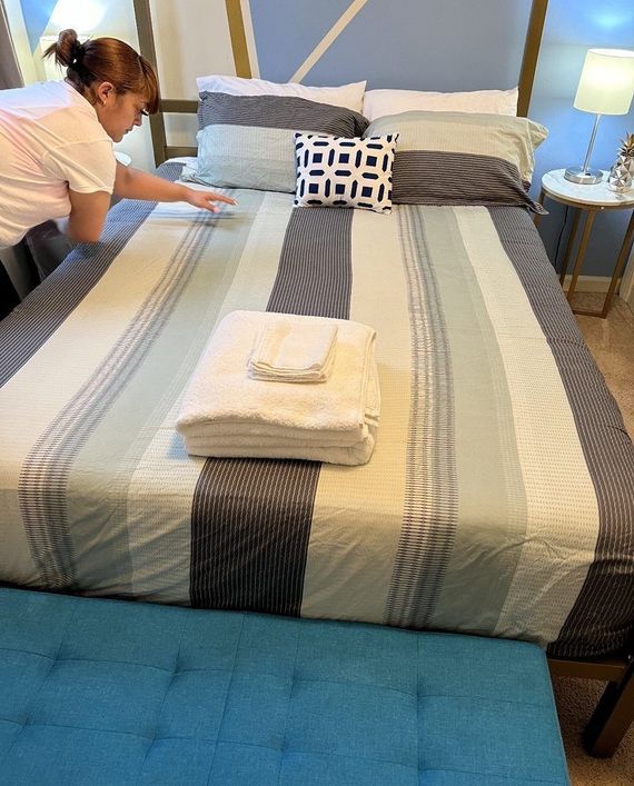 Person arranging a bed with striped comforter and a blue bench at the foot. White towels are stacked on the bed.